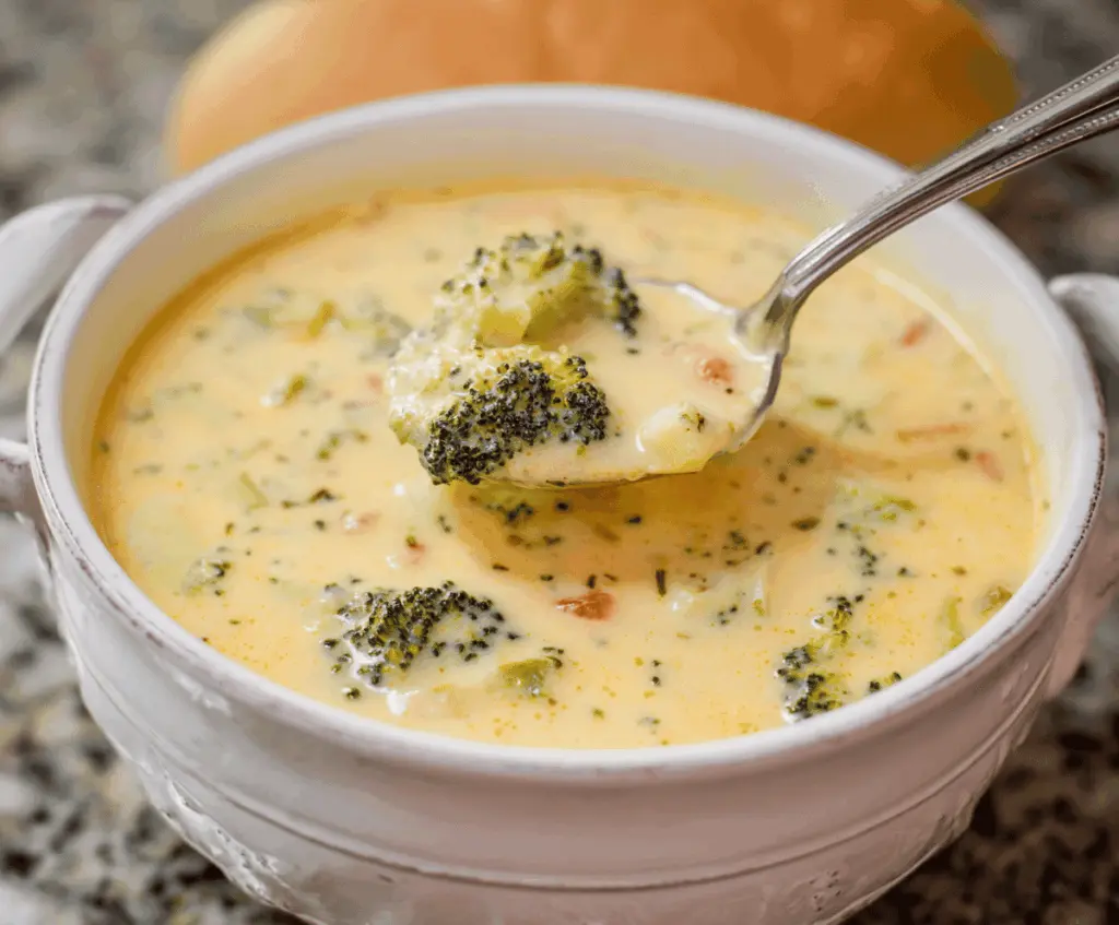 A bowl of creamy potato broccoli cheddar soup garnished with shredded cheese and fresh herbs, served with crusty bread on a rustic wooden table.
